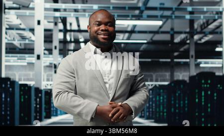 Smiling manager in data center doing checkup on hardware to avoid downtime. Happy african american supervisor overseeing server room infrastructure supercomputers, camera B Stock Photo
