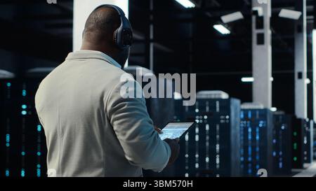 Admin walking in data center, listening music while doing inspection on infrastructure tech. IT specialist in server hub wearing headphones analyzing traffic patterns of equipment mainframes, camera B Stock Photo
