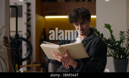 Concentrated Hispanic guy reading literature holding book at home hipster turning page focused smart adult man reader male looking text poetry Stock Photo