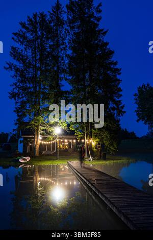 A cozy lakeside cabin in rural Estonia lit by warm outdoor lights, reflected in calm water on a peaceful summer night. Stock Photo