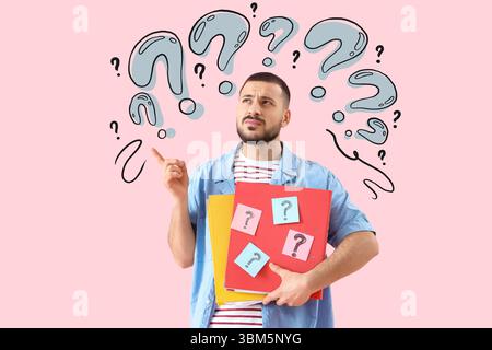 Portrait of thoughtful young man holding folders with question marks on pink background Stock Photo