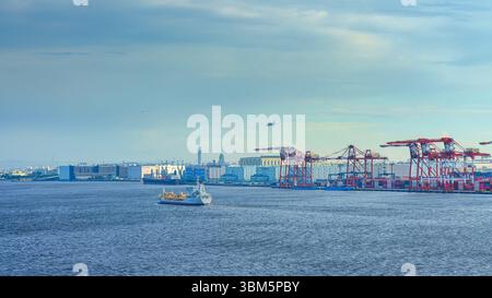 Tokyo, Japan - Oct 2 2024, Panoramic view of docks of Tokyo port with many cargo cranes lined up, with small boat sailing in foreground, with a plane Stock Photo