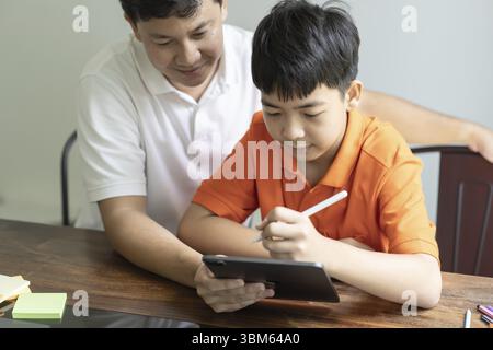 Asian father and son using tablet teaching son to draw playing with digital tablet at desk fatherhood concept Stock Photo
