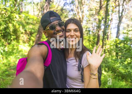 Interracial couple smiling and waving while taking a selfie during a vibrant hike in a lush, green forest filled with sunlight Stock Photo