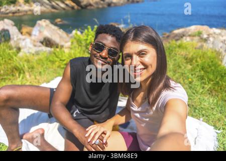 Young multiracial couple holding hands and taking a selfie while enjoying a sunny day by the sea Stock Photo