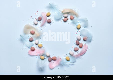 Happy easter greeting card. Round frame made of colorful feathers and chocolate eggs in sugar glaze on pastel blue background. Flat lay, copy space Stock Photo