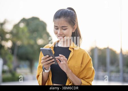 Smiling young Asian woman using smartphone in urban city checking apps mobile outdoors Stock Photo