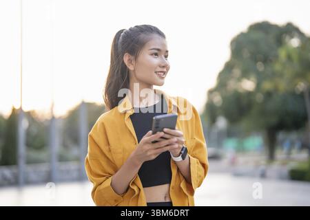 Smiling young Asian woman using smartphone in urban city checking apps mobile outdoors Stock Photo