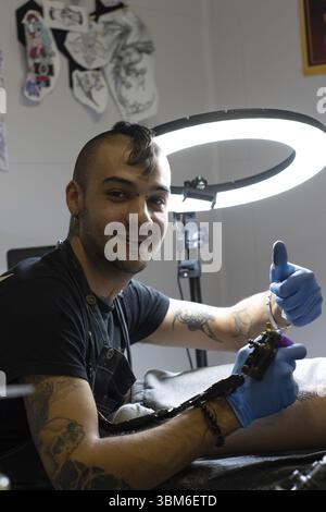 Young tattoo artist performing the OK gesture while tattooing in his tattoo studio Stock Photo