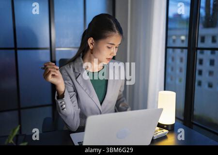 Alone Working Late at Night. Asian business woman at workplace in office late night work at a desk in a dark office working on a laptop late in the ev Stock Photo