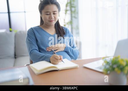 Pregnant asian woman writing notes and using a laptop while working at home pregnant businesswoman working on remote project in home office pregnancy Stock Photo