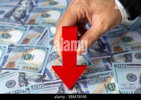 A hand holds a red arrow pointing downwards against a background of scattered US one-hundred dollar bills. Financial downturn, economic loss Stock Photo