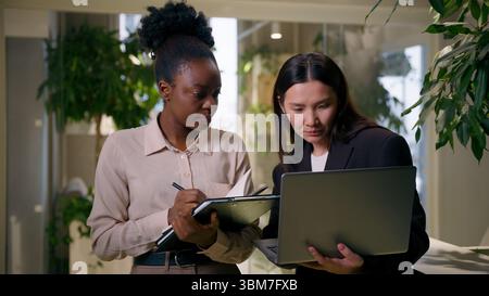 African Asian business women girls colleagues multiracial businesswomen in office teamwork laptop project writing tasks notebook Chinese Korean Stock Photo