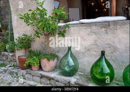 Several large, green, bulbous glass bottles stand on a low, old wall next to a house wall with some flowerpots and green plants Stock Photo