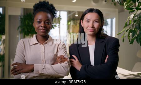 Two confident businesswomen multiracial diverse African Asian girls portrait women in office posing together crossed arms Chinese American business Stock Photo