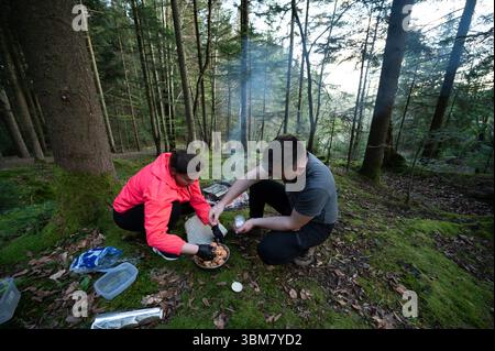Two friends enjoy cooking over an open fire in a tranquil forest, gathering ingredients and savoring the moment amidst nature's beauty and fresh air. Stock Photo