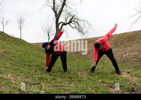 Two individuals in red jackets stretch on a grassy slope under a cloudy sky, embracing a healthy morning routine surrounded by nature's tranquility du Stock Photo