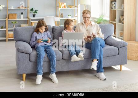 Smiling grandmother sitting on sofa with grandchildren using devices and spending time together. Stock Photo