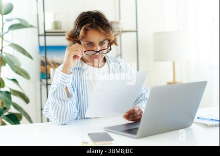 Serious brazilian successful businesswoman with glassed reading printed report while working on laptop at home. Focused professional analyzing data or financial charts during remote work in apartment Stock Photo