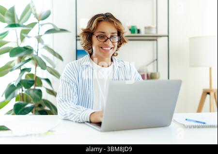 Happy brazilian woman freelancer using laptop while working from her modern apartment. Confident young woman engaged in remote work or online communication in home office environment Stock Photo