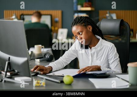 African American woman working in modern office, talking on smartphone and using computer at desk, displaying multitasking and concentration Stock Photo