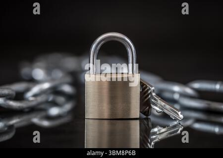 Padlock and key on reflective surface in dark studio setting, with blurred chain in background on black. Close-up product photo. Stock Photo
