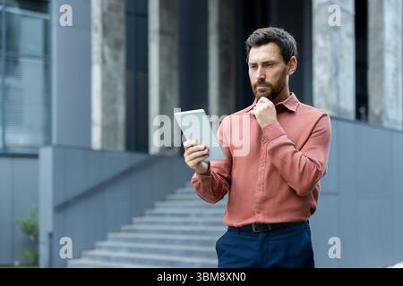 A thoughtful businessman in a red shirt is looking at his tablet in an office setting. He is touching his chin while pondering. Stock Photo