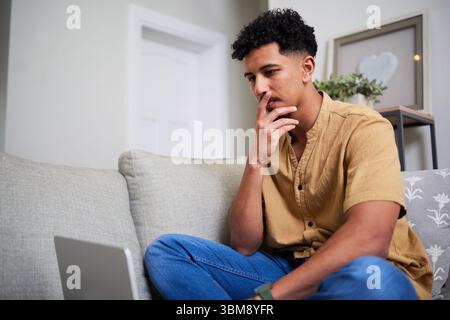 Thoughtful Young Man Sitting on a Couch, Reflectively Looking at a Laptop Screen in a Modern Living Room Stock Photo