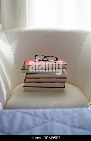 a pile of hardback books and eyeglasses on a white armchair Stock Photo
