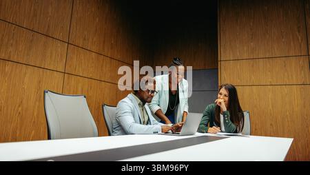 A corporate team is gathered around a boardroom table, engaged in a meeting. The setting is professional, with three colleagues collaborating over a l Stock Photo