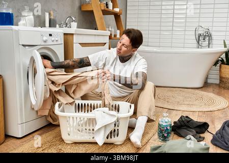 Tattooed young man blends comfort and household chores while doing laundry at his home. Stock Photo