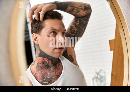 Tattooed man takes a moment to adjust his hair in a serene home environment. Stock Photo