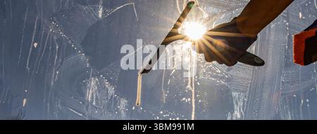 Woman's hand in rubber glove cleaning window with a squeegee on a sunny day. Sunlight, soap streaks, and cleaning tools create a realistic scene of sp Stock Photo