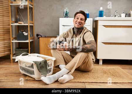 A tattooed young man smiles on the floor, using his phone in a cozy home. Stock Photo