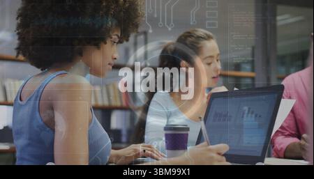 Working curly-haired woman typing on laptop in library office, with printed charts, pen, coffee cup. Collaboration, teamwork, data analysis, professio Stock Photo