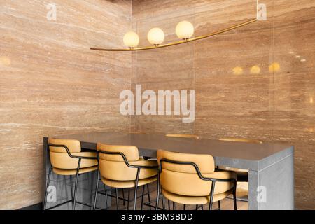 A luxury dining room detail with orange granite slab walls, a gold chandelier, and leather bar stools sitting at a grey stone table. Stock Photo