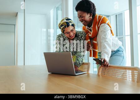 Two diverse women are joyfully collaborating in a stylish office environment, using a laptop to discuss creative ideas. Teamwork, modern business dyna Stock Photo