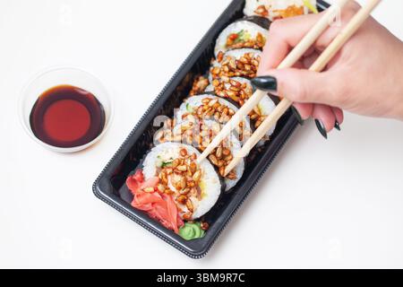 Woman holding sushi roll. Chopsticks grip traditional rice roll. Asian lunch with soy sauce in small bowl. Japanese cuisine takeaway meal. Stock Photo
