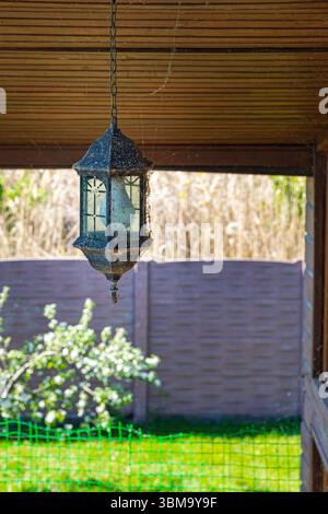 Rusty hanging lantern with cobwebs under wooden canopy in garden. Symbol of neglect, rural life, or seasonal cleaning with spring sunlight and backyar Stock Photo