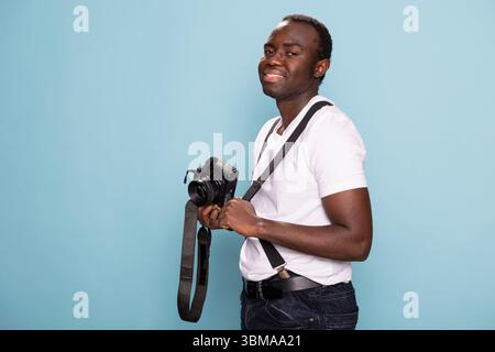 African american man stands confidently, holding a camera in one hand while adjusting his suspenders strap with the other. Portrait of black male model in casual attire, posing with his DSLR. Stock Photo