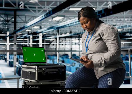 Mockup laptop next to female engineer in server hub using tablet to optimize hardware performance. African american woman in data center using green screen notebook, upgrading rackmounts Stock Photo