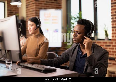 Black male entrepreneur wearing a headset, attentively listening during an online marketing presentation. African American wheelchair user communicating with clients in a brick walled office. Stock Photo