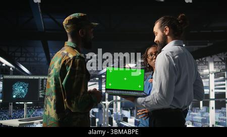 Army tech specialist in data center and coworkers using green screen laptop to troubleshoot military computer systems. Soldier and command center server room admins using chroma key notebook, Stock Photo