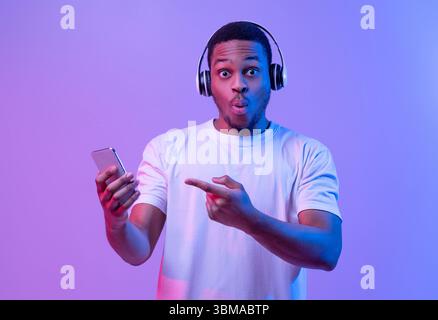 Cool App. Surprised Black Guy In Wireless Headphones Pointing At Smartphone Stock Photo