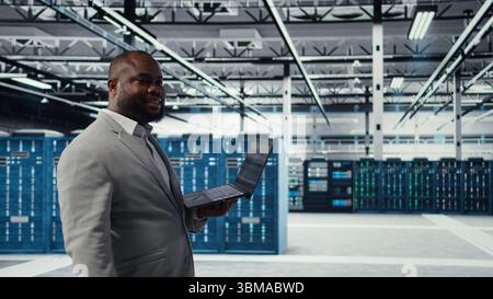 Data center engineer deploying AI driven management tools using laptop. Server farm employee inspecting server clusters, updating network topologies using artificial intelligence, camera A Stock Photo