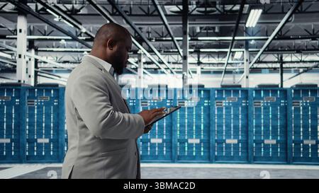 Cheerful server room engineer troubleshooting backend operations using software on tablet. Jolly data center programmer compiling software patches to fix bugs using device, camera A Stock Photo
