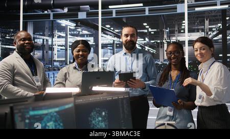 Smiling engineering team in server room reviewing infrastructure diagnostics on devices. IT professionals in data center analyzing performance data to maintain system integrity and uptime, camera A Stock Photo