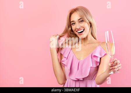Portrait of happy young woman holding glass of sparkly champagne, smiling and looking at camera on pink background Stock Photo