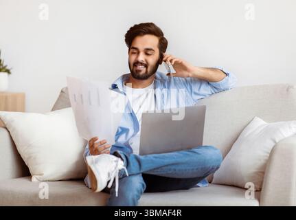 Eastern Freelancer Man With Cellphone, Papers And Laptop Working Remotely At Home Stock Photo