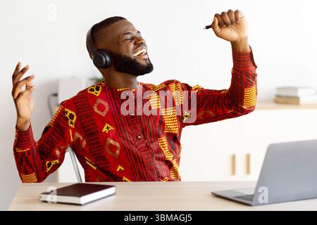 Joyful african freelancer man having fun at workplace, singing and listening music Stock Photo